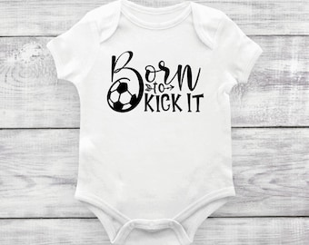May include: A white short-sleeved baby bodysuit featuring a black graphic print of a soccer ball and the text Born to KICK IT. The design includes a small arrow icon between the words to and KICK, with the soccer ball replacing the letter O.