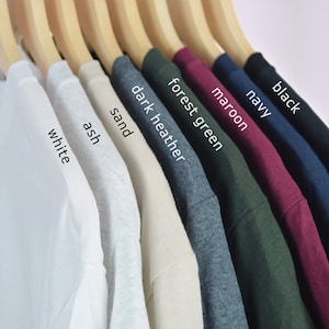 Może przedstawiać: Rząd dziesięciu koszulek wiszących na drewnianych wieszakach. Koszulki są w następujących kolorach: biały, popielaty, piaskowy, ciemnoszary melanż, zielony leśny, bordowy, granatowy, czarny.