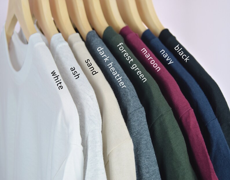 Może przedstawiać: Rząd dziesięciu koszulek wiszących na drewnianych wieszakach. Koszulki są w następujących kolorach: biały, popielaty, piaskowy, ciemnoszary melanż, zielony leśny, bordowy, granatowy, czarny.