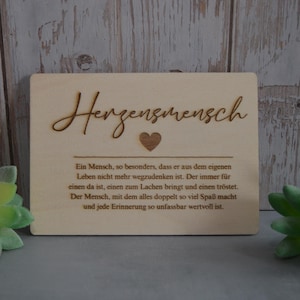 May include: A light brown wooden plaque with the word "Herzensmensch" in cursive script, a heart symbol, and a paragraph of text. The plaque is rectangular, with a wood grain background and green succulent plants.