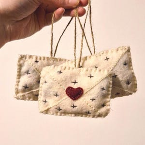 May include: Three cream-colored felt envelope ornaments with black cross stitch details and a red heart. Each ornament is suspended by a natural twine string, perfect for holiday or everyday decor.