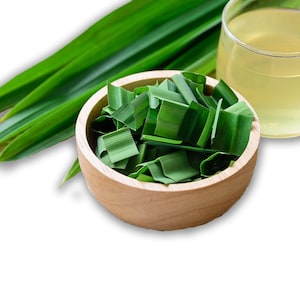 May include: A round wooden bowl filled with chopped green pandan leaves, accompanied by several long, whole pandan leaves and a clear glass containing a pale yellow liquid, all arranged together on a plain white background.