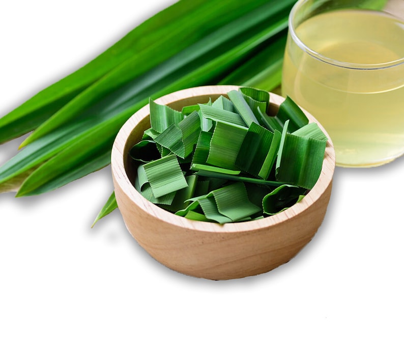 May include: A round wooden bowl filled with chopped green pandan leaves, accompanied by several long, whole pandan leaves and a clear glass containing a pale yellow liquid, all arranged together on a plain white background.