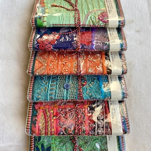 Könnte beinhalten: Ein Stapel von sechs handgefertigten Papiernotizbüchern, verpackt in durchsichtigem Kunststoff. Jedes Notizbuch hat ein einzigartiges Patchwork-Design mit aufwendiger Stickerei, Pailletten und Perlen. Die Notizbücher sind mit einer rot-braunen Schnur gebunden. Der Text "HAND MADE PAPER NOTE BOOK" ist sichtbar.