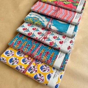 May include: Six notebooks with patterned fabric covers in a stack, wrapped in clear plastic. The covers display various designs in red, blue, yellow, and green. Each notebook is tied with twine and has a paper band that reads "HAND MADE".