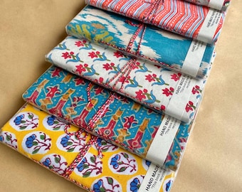 May include: Six notebooks with patterned fabric covers in a stack, wrapped in clear plastic. The covers display various designs in red, blue, yellow, and green. Each notebook is tied with twine and has a paper band that reads "HAND MADE".