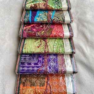 Può includere: Una pila di sei diari rettangolari avvolti in plastica trasparente, ognuno caratterizzato da una copertina in tessuto unica con ricami complessi, paillettes e perline in colori vivaci tra cui verde, blu, viola, rosa e arancione, legati con spago.