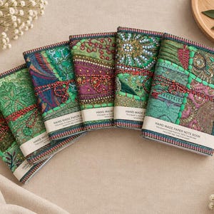 May include: Five rectangular notebooks arranged in a fan shape, each featuring a unique fabric cover with intricate embroidery, beadwork, and sequins in shades of green, purple, and teal. Each notebook has a white paper band labelled HAND MADE PAPER NOTE BOOK.