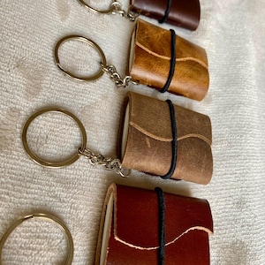 Op de afbeelding: Vier kleine leren notitieboekjes-sleutelhangers in bruine tinten, vastgezet met zwarte elastiekjes. Elke sleutelhanger heeft een zilverkleurige ring. De notitieboekjes liggen op een wit, getextureerd oppervlak.