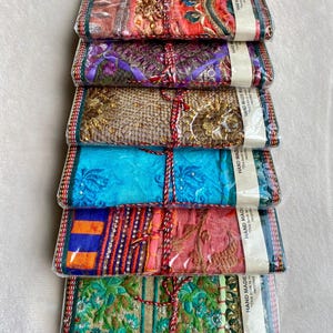 May include: A stack of six colorful, rectangular notebooks wrapped in clear plastic. Each notebook features a unique fabric design with intricate embroidery and embellishments. The notebooks are tied with red and white twine.