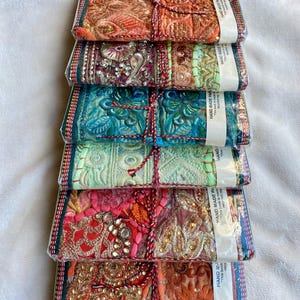 May include: Six hand-made paper notebooks, each wrapped in clear plastic and tied with red and white twine. The notebooks display detailed embroidery in orange, teal, pink, and gold. The text on the label reads "HAND MADE PAPER NOTEBOOK".