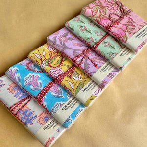 Puede incluir: Una pila de seis cuadernos con estampados florales en varios colores, incluyendo rosa, azul, amarillo y morado. Cada cuaderno está envuelto y atado con hilo rojo. El texto "HAND MADE PAPER NOTE" es visible en la etiqueta.
