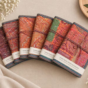 May include: Six rectangular notebooks with fabric covers featuring intricate, colourful embroidered patterns in shades of red, orange, green, and purple. Each notebook is secured with a thin cord and has a white paper band labelled HAND MADE PAPER NOTEBOOK.