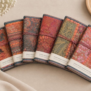 May include: Six rectangular notebooks with fabric covers featuring intricate floral and paisley patterns in shades of orange, red, green, and gold. Each notebook is secured with a thin cord and has a white paper band labelled HAND MADE PAPER NOTEBOOK.