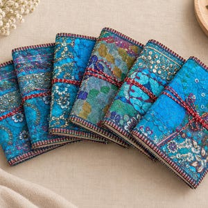 May include: Five rectangular journals with fabric covers in shades of blue, teal, and green. Each cover features intricate embroidery, sequins, and floral patterns, secured with a thin red and white twisted cord tied in a bow around the centre.