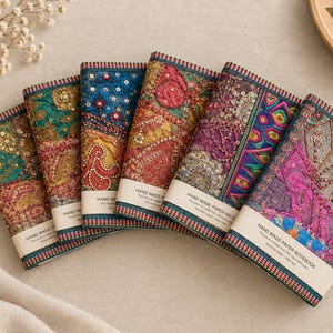 May include: Six rectangular notebooks arranged in a fan shape, each featuring a unique fabric cover with intricate embroidery, sequins, and beadwork in vibrant colours like teal, gold, blue, red, and purple, with a paper band labelled HAND MADE PAPER NOTEBOOK.