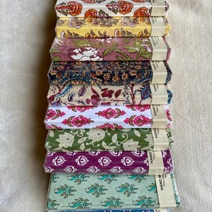May include: A collection of notebooks with floral designs in various colours. The notebooks are wrapped in clear plastic and feature patterns in shades of orange, yellow, purple, and blue. The packaging includes the text "HAND MADE PAPER NOTEBOOK".