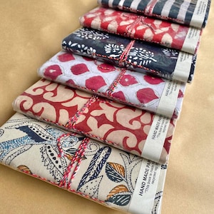 May include: A collection of handmade paper journals with fabric covers. The journals display diverse patterns, such as red and white florals, blue and beige paisley, and striped designs. Each journal is bound with red and white twine.