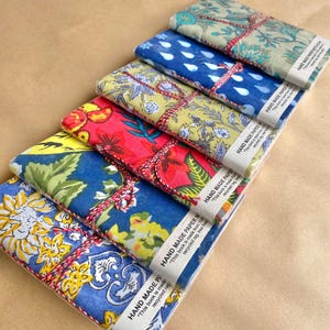 May include: A selection of handmade paper notebooks, each covered in a different floral fabric design. The notebooks are tied with red and white twine and labelled "HAND MADE PAPER NOTE". The fabric patterns feature blue, yellow, red, and green hues.