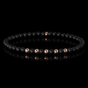 Puede incluir: Un brazalete de cuentas negras con detalles en oro rosa. El brazalete está hecho de cuentas negras mate con un acabado liso. Los detalles en oro rosa son cuentas pequeñas y redondas que están espaciadas uniformemente en todo el brazalete.