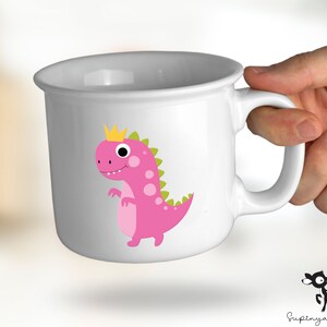 K&ouml;nnte beinhalten: Wei&szlig;e Keramiktasse mit einem rosa Dinosaurier, der eine goldene Krone tr&auml;gt. Der Dinosaurier blickt nach links und hat gr&uuml;ne Flecken. Die Tasse hat einen Henkel.