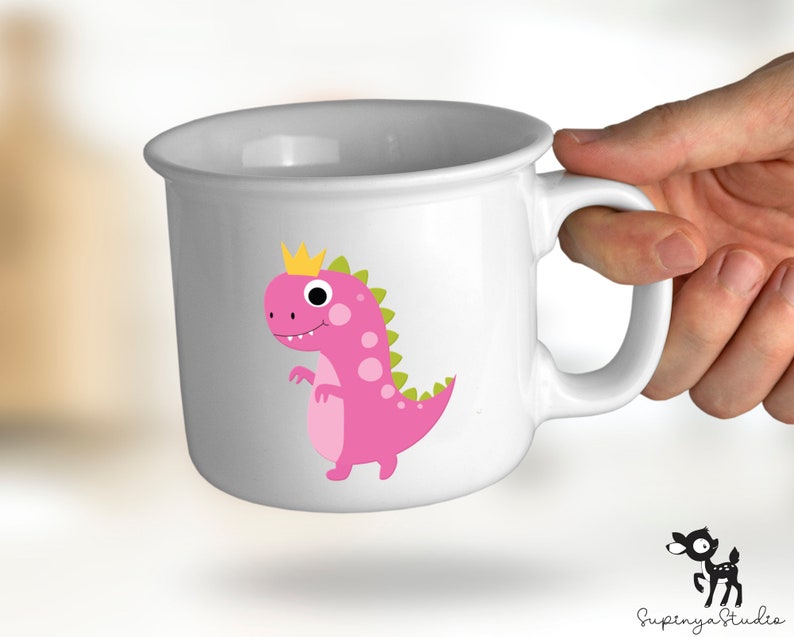 K&ouml;nnte beinhalten: Wei&szlig;e Keramiktasse mit einem rosa Dinosaurier, der eine goldene Krone tr&auml;gt. Der Dinosaurier blickt nach links und hat gr&uuml;ne Flecken. Die Tasse hat einen Henkel.