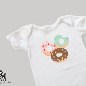 May include: White baby onesie featuring a design of three donuts. The donuts are pink with hearts, green with sprinkles, and brown with chocolate icing and sprinkles.