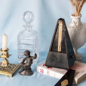 Könnte beinhalten: Eine Stillleben mit einem Messing-Kerzenleuchter mit einer weißen Kerze, einer klaren Glaskaraffe, einer kleinen Bronzestatue eines Cherubs, einem schwarzen Metronom, einer weißen Porzellanvase mit getrockneten Weizenhalmen und einem Buch mit dem Titel "La Sine" auf einem hellblauen Hintergrund.