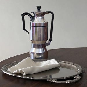 May include: A silver-toned metal coffee percolator with a black handle and lid knob, positioned behind an oval silver-toned metal serving tray. A folded white cloth napkin rests on the surface of the tray, which features decorative handles.