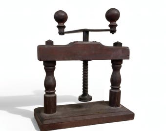 May include: A vintage wooden press with a dark brown finish. The press features a screw mechanism, two decorative turned legs, and a handle with two spherical knobs. The base is rectangular, and the overall design suggests a tool for pressing or clamping.