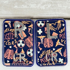 Puede incluir: Dos portatarjetas rectangulares azul marino con temática médica. El carrete izquierdo tiene las letras "R" y "CR" en plata, con las palabras "radiate positive vibes". El carrete derecho tiene las letras "L" y "CR" en plata, con las palabras "radiate positive vibes".