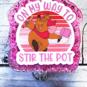 May include: Pink glitter badge holder with a cartoon mouse holding a spoon and a pot. The text on the badge reads "On my way to stir the pot."
