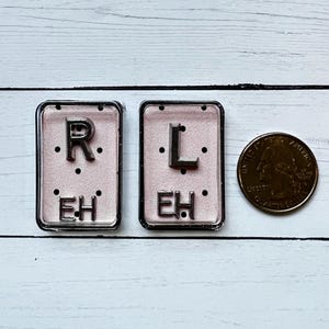 May include: Two rectangular, silver-framed name tags with the letters "R" and "L" above "EH" on a pink background. A US quarter coin is next to the name tags. The name tags are on a white wooden surface.