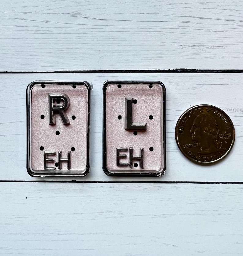 May include: Two rectangular, silver-framed name tags with the letters "R" and "L" above "EH" on a pink background. A US quarter coin is next to the name tags. The name tags are on a white wooden surface.