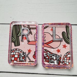 May include: Two rectangular resin pieces with pink backgrounds featuring a cowboy hat, cactus, disco ball, and stars. One has the letter "R" and "EK", the other has "L" and "EK". The edges are glittery.