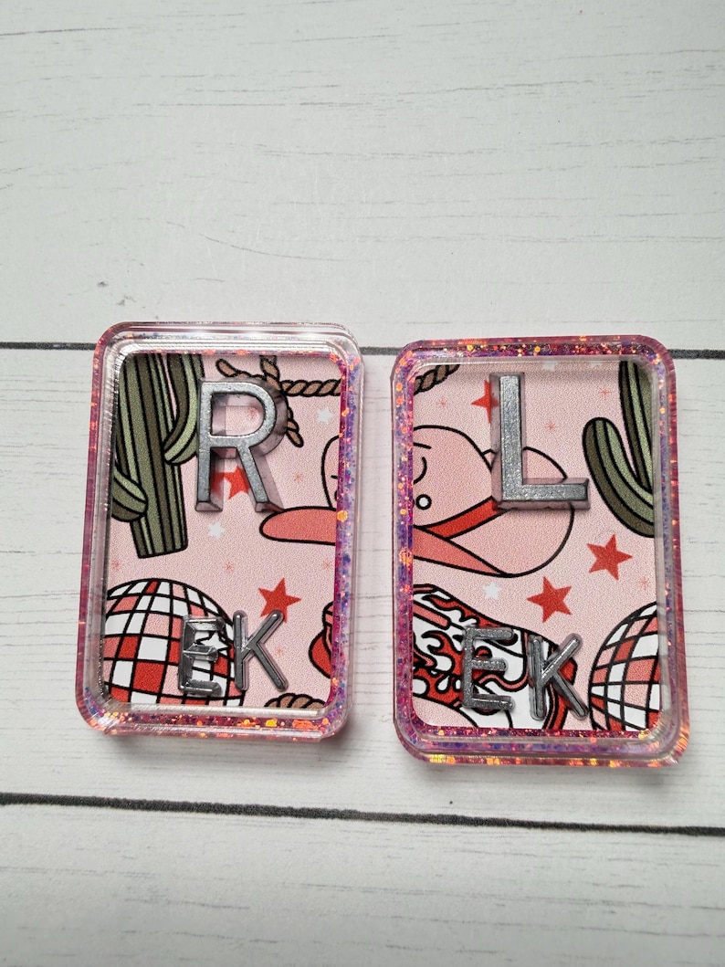 May include: Two rectangular resin pieces with pink backgrounds featuring a cowboy hat, cactus, disco ball, and stars. One has the letter "R" and "EK", the other has "L" and "EK". The edges are glittery.