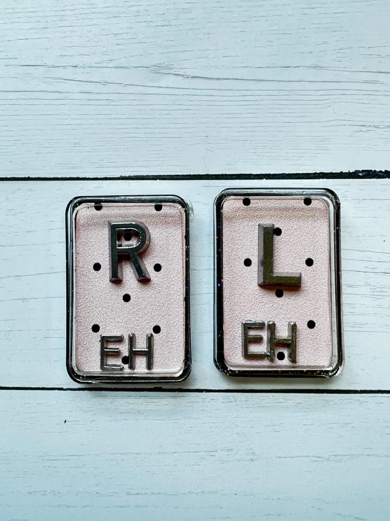 May include: Two rectangular, clear-framed accessories with a pink background. The left one has the letter "R" above the letters "EH". The right one has the letter "L" above the letters "EH".