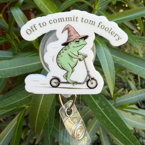 May include: A badge reel featuring a green frog wearing a wizard hat and riding a scooter. The text "Off to commit tom foolery" is above the frog. The reel has a clear plastic ID holder and a metal clip.