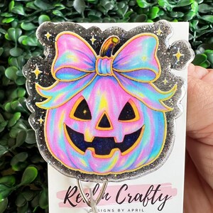 May include: A colorful badge reel featuring a jack-o'-lantern design with a large bow. The pumpkin is pink, blue, and yellow with a black face. The edges are outlined with black glitter and stars. The text "Reach Crafty" is visible.
