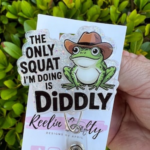 May include: A clear acrylic badge reel with a glittery border and a cartoon frog wearing a cowboy hat. The text reads "The only squat I'm doing is Diddly." The badge reel has a metal clip and a clear plastic strap.