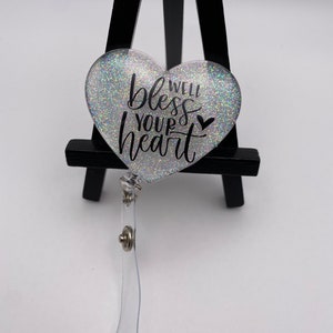 May include: A silver glitter heart-shaped badge holder with a black inscription that reads "Well bless your heart" with a small heart symbol.