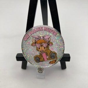 May include: A white glitter badge holder with a cartoon cow wearing sunglasses and a pink bandana. The text "One Boujee Heifer" is printed on the badge holder.