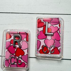 May include: Two rectangular keychains with a pink heart pattern. One keychain has a silver "R" and "CC" charms, the other has a silver "L" and "CC" charms. The keychains have a clear, glittery border.