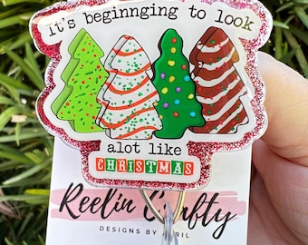 Op de afbeelding: Een rode glitter badgehouder met een witte achtergrond. De badgehouder heeft een ontwerp met drie kerstboom met de tekst "It's beginning to look a lot like Christmas". De tekst "Reelin' & Crafty Designs by RIL" is onder het ontwerp gedrukt. Social media-iconen voor TikTok, Instagram en Facebook zijn onder de tekst gedrukt.