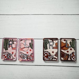 May include: Four rectangular resin keychains. Two keychains have a pink background with a cowboy hat, cactus, and disco ball pattern. The other two have a brown background with a cowboy hat, cactus, and disco ball pattern. Each keychain has silver letters.