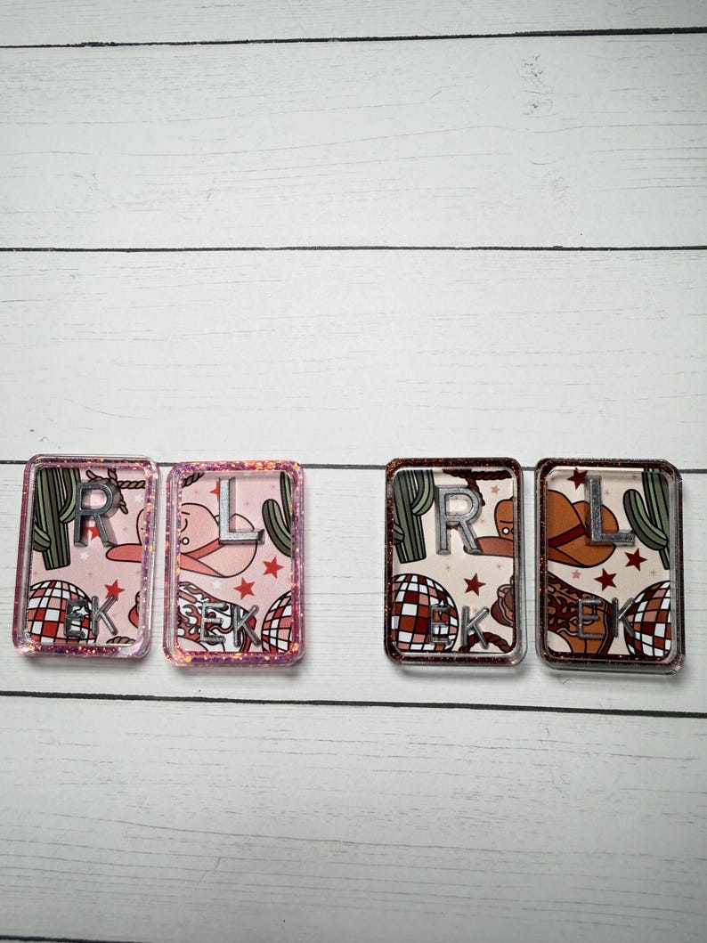 May include: Four rectangular resin keychains. Two keychains have a pink background with a cowboy hat, cactus, and disco ball pattern. The other two have a brown background with a cowboy hat, cactus, and disco ball pattern. Each keychain has silver letters.