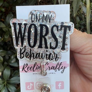 May include: A badge reel with the text "ON MY WORST Behavior" in black lettering. The badge reel has a clear, glittery background and a silver metal clip. The card below the reel has the text "Reelin Crafty" and social media icons.