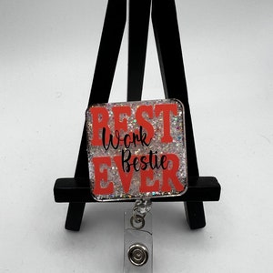 Puede incluir: Un portagafete transparente con un clip plateado y un encanto cuadrado de resina naranja y blanca brillante que dice "Best Work Bestie Ever".