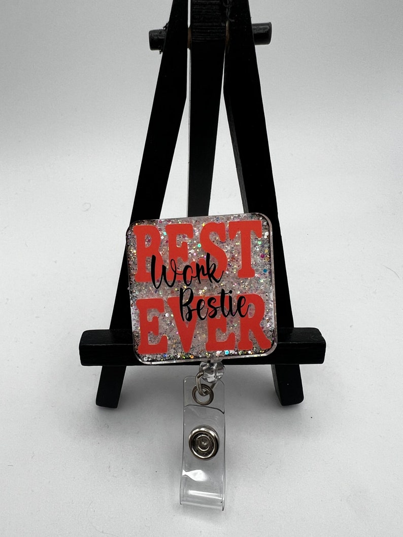 Puede incluir: Un portagafete transparente con un clip plateado y un encanto cuadrado de resina naranja y blanca brillante que dice "Best Work Bestie Ever".