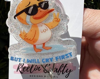 May include: A white glitter badge holder with a cartoon duck wearing sunglasses and a peace sign. The duck is crying and the text "I will handle it but I will cry first" is written on the holder. The holder is attached to a silver retractable badge reel with the text "Reelin & Crafty Designs by RIL" on the bottom.